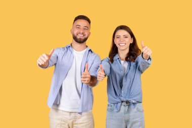 İki gülümseyen yetişkin, erkek ve kadın, sarı bir arka planda başparmak hareketi yaparak, pozitiflik ve onayı ifade ediyorlar.