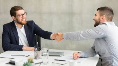 Two professionally dressed businessmen in a modern office environment sealing a deal with a firm handshake over a meeting table with documents clipart