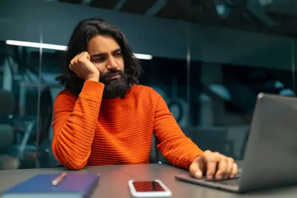 Middle-aged man in an orange sweater appears bored while using a computer at a desk