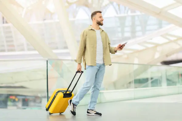 空港で携帯電話を使用しながら明るい黄色のスーツケースで散歩するファッショナブルな男 — ストック写真
