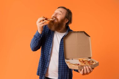 Kafkasyalı kızıl sakallı adam elinde karton teslimat kutusuyla pizza dilimi yerken turuncu arka planla stüdyoda dikiliyor. Konforlu yemek yeme ve aldatma konsepti