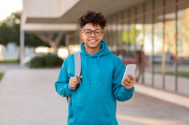 A cheerful student brazilian guy with stylish glasses and headphones around neck holds a smartphone, urban background clipart