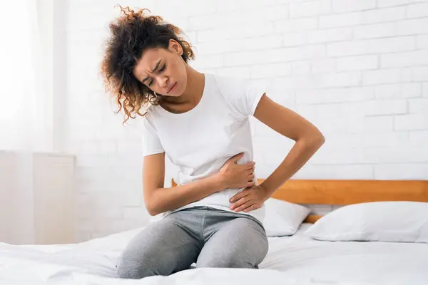 stock image African-american girl suffering from abdominal pain, sitting on bed at home