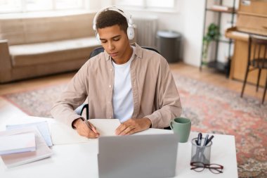 Kulaklığı olan odaklanmış genç bir erkek, yanında laptop, kahve fincanı ve gözlüklerle ahşap bir masada kitap okuyor.