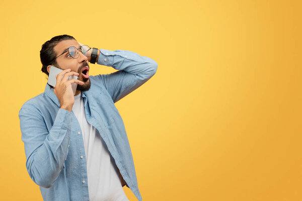 Озадаченный молодой человек в джинсовой рубашке, разговаривающий по телефону и трогающий голову в замешательстве или забывчивости на ярко-желтом фоне