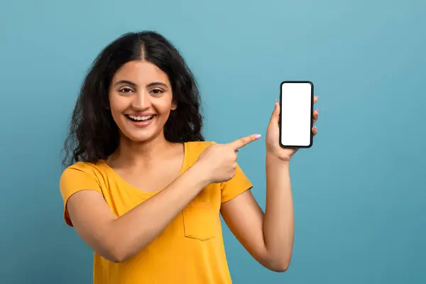 一个快乐的女人指着一个空白的智能手机屏幕 在沉闷的背景上带着大大的笑容 — 图库照片