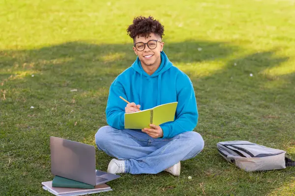 眼鏡をかけた若いブラジル人男性がノートブックに書き込み ラップトップとバックパックで草の上に座りながら — ストック写真