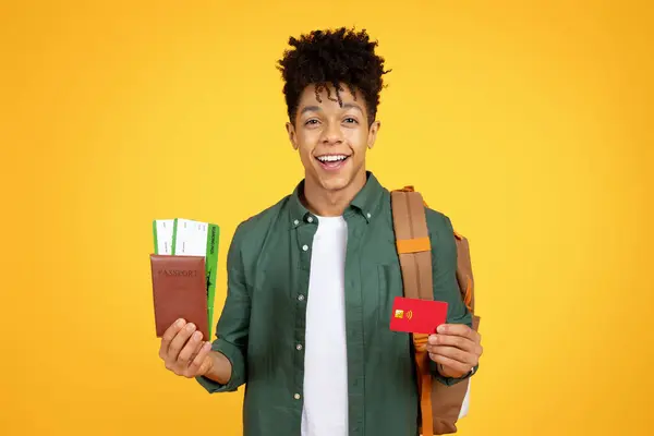 バックパックを着用している自信のある若いアフリカ系アメリカ人男性は 黄色で旅行する準備ができていることを表すパスポートとクレジットカードを持っています — ストック写真