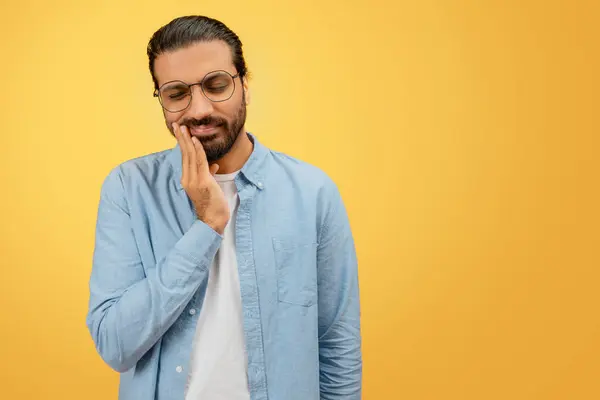 一个戴眼镜的不舒服的印地安人在黄色的统一背景下托着他的面颊 暗示着牙痛或不适 — 图库照片