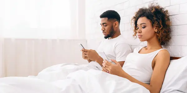 アフリカ系アメリカ人の若いカップルはベッドに戻り スマートフォンに囲まれ 現代の関係の問題を示しています — ストック写真