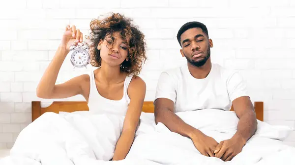 We should go to bed earlier. Sleepy african-american couple sitting in bed with alarm clock, cant wake up in morning, copy space
