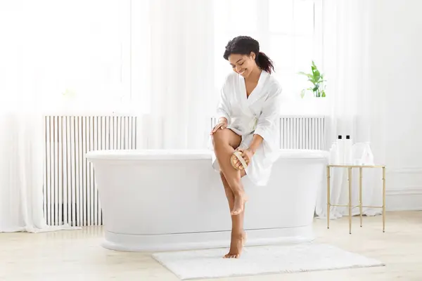 一位快乐的非洲裔美国女人 身穿长袍 坐在明亮 干净的浴室里 用刷子按摩她的腿 — 图库照片