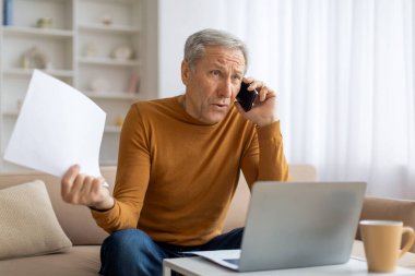 Bilgisayarı kullanırken belgelerle telefonda kıdemli bir adam arıyor, belki işle ya da mali durumla ilgileniyordur.