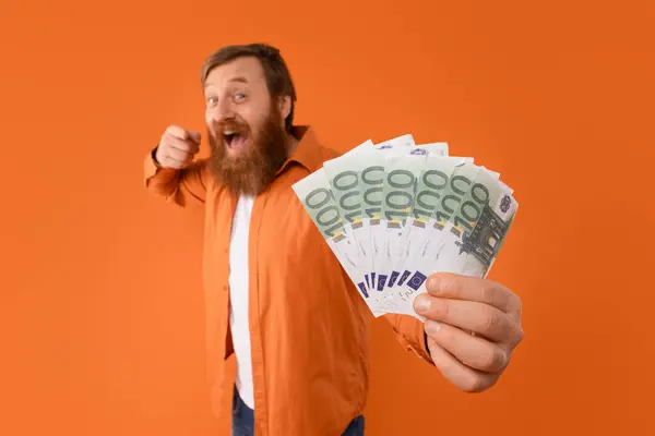 Money for you. Happy redhaired man with beard holds handful of Euro cash, shouting in celebration of financial success, pointing finger at camera over orange studio backdrop. Selective focus
