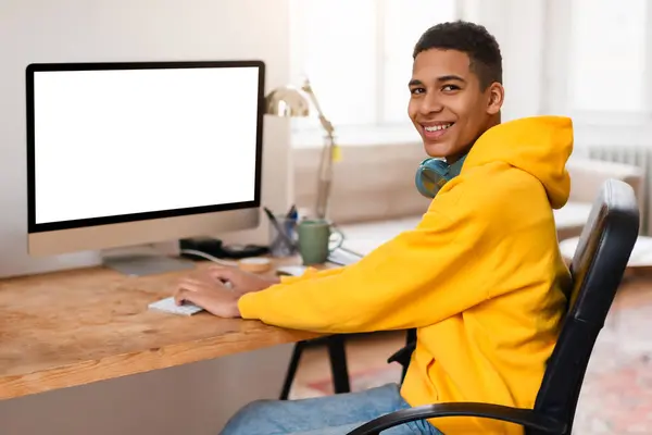 明るい部屋のカメラで空白のスクリーンと笑顔のコンピュータを使用して ヘッドフォンが付いている黄色いパーカーの陽気なティーンエイジャーの男 — ストック写真