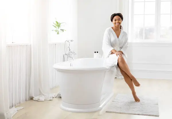 非洲裔美国妇女在一个干净 充满阳光的浴室里 坐在浴缸边 面带微笑 — 图库照片