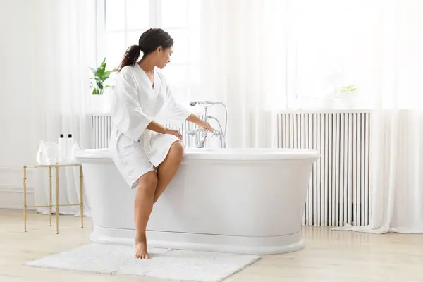身穿白色长袍的非洲裔美国妇女准备在灯光明亮 宁静的浴室里洗澡 — 图库照片