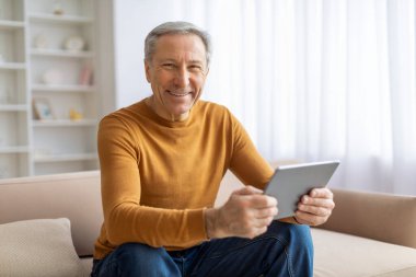 Neşeli bir gülümsemesi olan yaşlı bir adam rahat bir evde dijital tablet ekranına bakıyor.