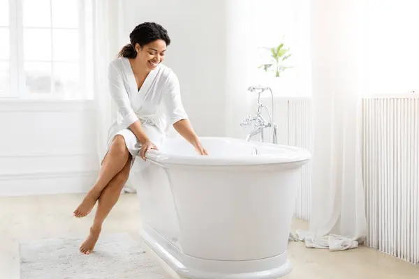 身穿白色长袍的快乐的非洲裔美国女人坐在整洁的浴室里独立的浴缸上 — 图库照片
