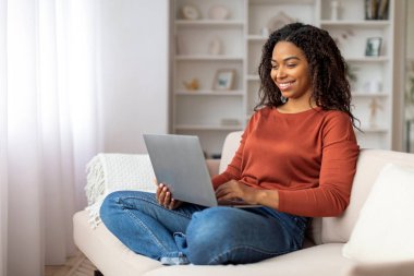 Günlük kıyafetli siyahi bir kadın kucağına dizüstü bilgisayarını yerleştirirken bir koltukta oturuyor.