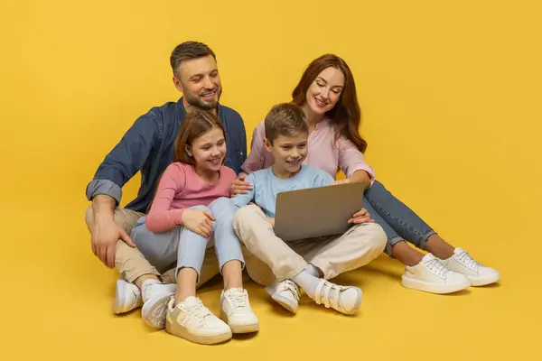 床に座っている笑顔の家族は 活気に満ちた黄色の背景で ラップトップと交流し 従事し 幸せです — ストック写真