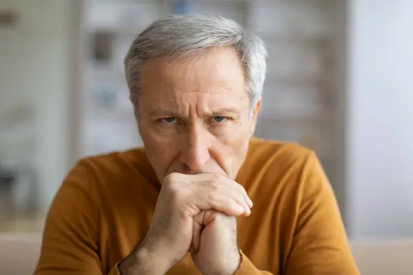 A contemplative senior man with hands clasped appears to be deep in thought or concern, home interior