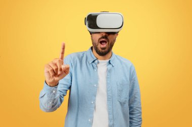 Indian man in casual attire is engaged with a virtual reality headset, pointing upwards, on a mustard background clipart