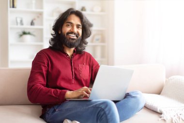 Dost canlısı bir tavrı olan rahat bir Hintli, dizüstü bilgisayarla çalışıyor, parlak bir odada koltukta rahatça oturuyor.