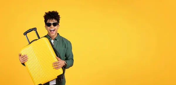 一个戴着太阳镜的友善快乐的非洲裔美国人拥抱着一个明亮的黄色行李箱 展示了在黄色背景下旅行的快乐和爱心 — 图库照片