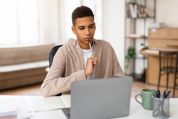 Задумчивый молодой человек с рукой на подбородке смотрит на экран ноутбука, глубоко в мыслях, возможно, решения проблем или изучения