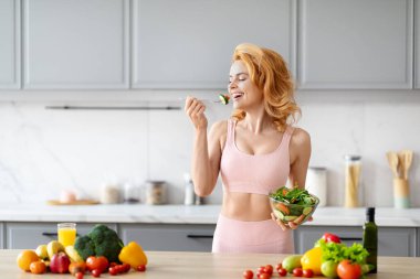 Altın saçlı çekici bir kadın modern mutfak ortamında taze yeşil salatanın tadını çıkarıyor.