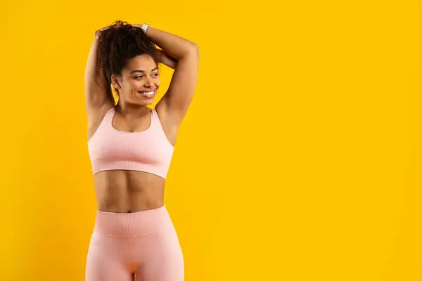 ピンクのアスレチックウェアの楽しいアフリカ系アメリカ人女性は 活気に満ちた黄色い背景に頭を向け 健康と活動を示しています — ストック写真