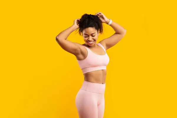 ピンクのトレーニング服の活気に満ちた若いアフリカ系アメリカ人女性は 黄色い壁に対するフィットネスセッションのために喜んで髪を準備します — ストック写真