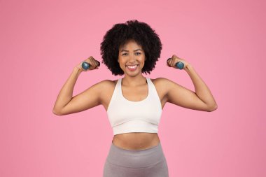 Doğal saçlı mutlu bir Afro-Amerikalı kadın, halterleri kaldırıyor, pembe arka planda spor yapıyor.