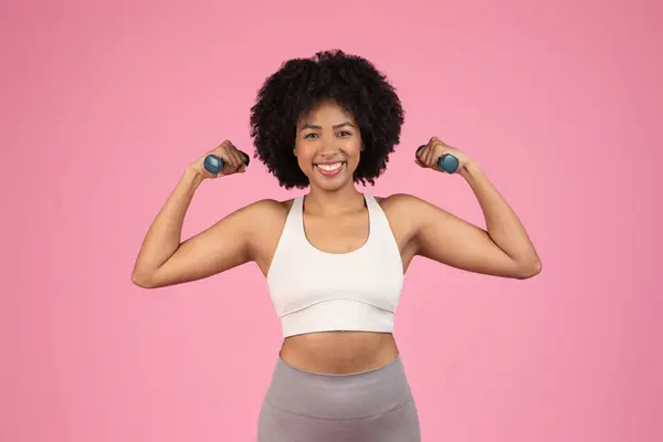 一位有着自然头发的快乐的非洲裔美国女人举起哑铃 在粉红的背景下展示出健康的身材 — 图库照片