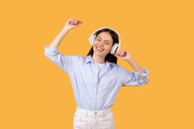 Cheerful woman with wireless headphones dancing and enjoying music, expressing happiness, yellow background clipart