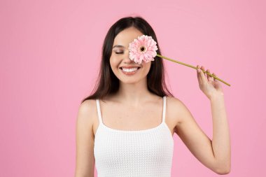 Neşeli genç bir kadın pembe bir Gerbera çiçeğini yüzünün yakınına yerleştirir.