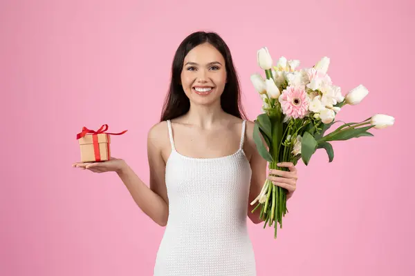 笑顔の若い女性は 柔らかいピンクの背景に設定された花束と小さな包まれた贈り物を提示します — ストック写真