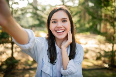 Güneşli bir parkta selfie çekerken gülümseyen neşeli kadın, mutluluğu ifade ediyor, güneşli günün tadını çıkarıyor.