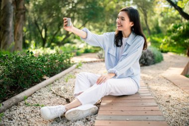 Enthusiastic woman capturing her own photo with a smartphone in a park setting depicts the joy of social sharing clipart