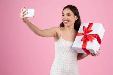 Beyaz elbiseli neşeli bir kadın pembe arka planda kırmızı kurdeleli büyük beyaz bir hediye kutusuyla selfie çeker.