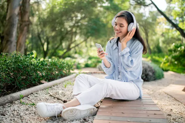 Beyaz Kulaklıklı Müzik Dinleyen Çevresi Yeşilliklerle Çevrili Bir Telefon Kullanan — Stok fotoğraf