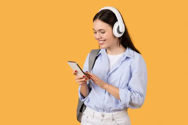 年轻女子带着耳机 专心致志地使用智能手机 连接在一起 观看黄色背景的播客 — 图库照片