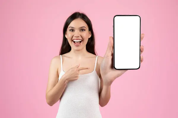 stock image Excited woman in white dress showcases smartphone with blank screen for advertising on pink background