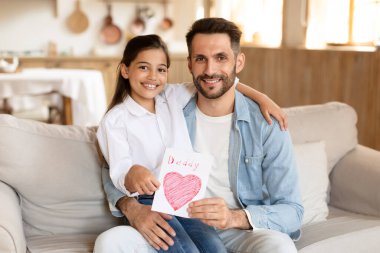 Gülümseyen baba ve küçük kızı kanepede el yapımı bir baba kartıyla oturuyor, sevgi ve neşe ifade ediyor.