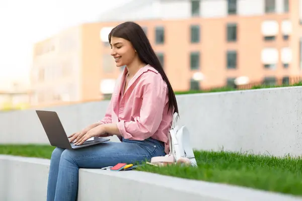 一个穿着休闲装的年轻姑娘坐在户外的窗台上 她使用笔记本电脑 看上去既满足又专注 — 图库照片