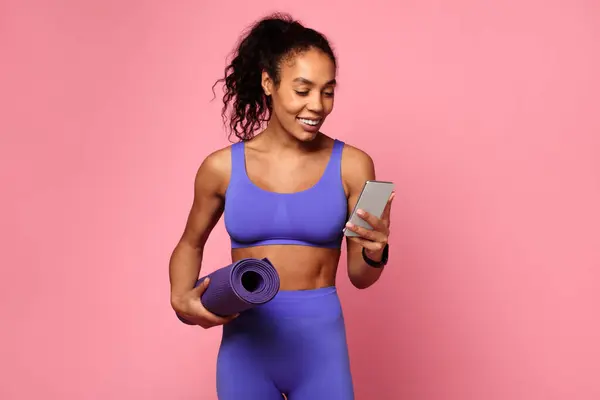 スポーツウェアのアフリカ系アメリカ人女性がヨガマットを持ち ピンクの背景でスマートフォンを見る — ストック写真
