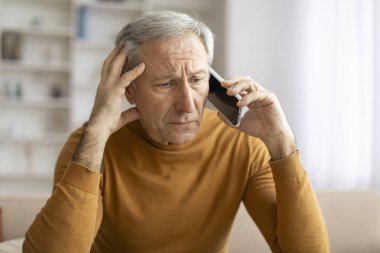 Endişeli yetişkin bir adam kulağına bir telefon tutar, endişeli ya da ev ortamında odaklanmış görünür.