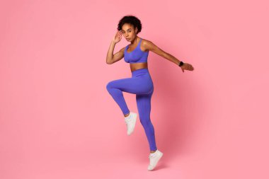 Canlı bir Afro-Amerikan kadın, mavi bir giysi giymiş, tek bacağı havada egzersiz yapıyor, pembe bir arka planda kendine güveni var.