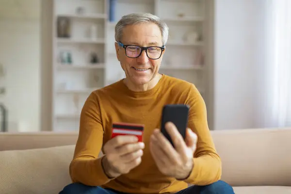 Cheerful senior man holding a smartphone and a credit card, possibly making a purchase from home